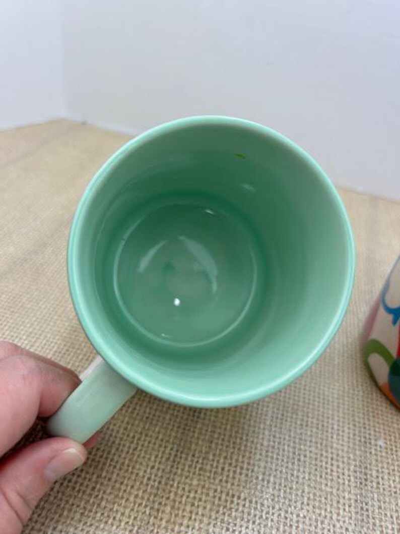 May include: A light green ceramic mug with a matching handle. The inside of the mug is also light green. The mug is sitting on a tan surface.