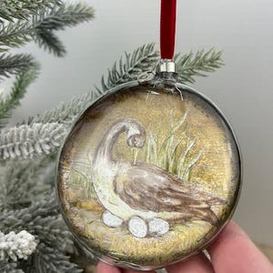 May include: A clear glass Christmas ornament with a hand-painted illustration of a goose and three eggs. The ornament has a red velvet ribbon for hanging. The background is a golden hue with green grass.