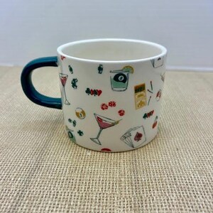 May include: White ceramic mug with a dark teal handle, decorated with colorful illustrations of cocktails, playing cards, and other bar-themed graphics. The mug is perfect for coffee, tea, or any beverage.