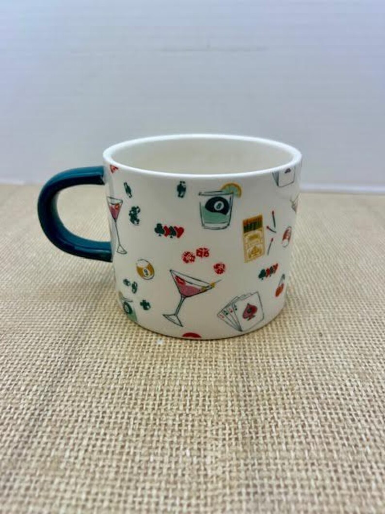 May include: White ceramic mug with a dark teal handle, decorated with colorful illustrations of cocktails, playing cards, and other bar-themed graphics. The mug is perfect for coffee, tea, or any beverage.