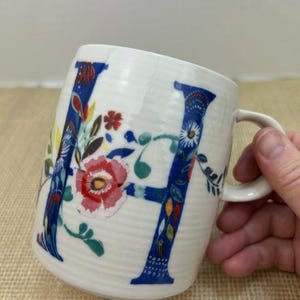 Puede incluir: Taza de cer&aacute;mica blanca con un dise&ntilde;o floral azul de la letra "H". La letra est&aacute; adornada con flores y hojas coloridas. La taza tiene un asa curva y una superficie texturizada.
