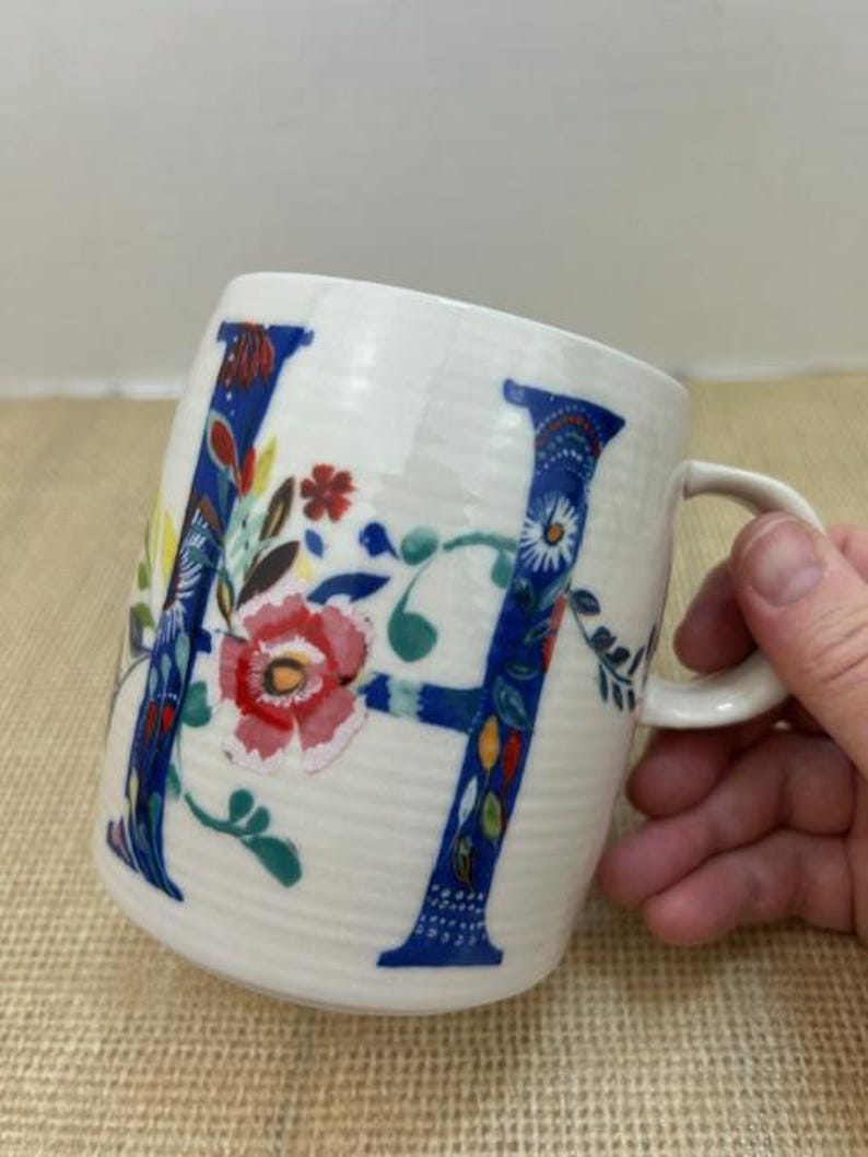 Puede incluir: Taza de cer&aacute;mica blanca con un dise&ntilde;o floral azul de la letra "H". La letra est&aacute; adornada con flores y hojas coloridas. La taza tiene un asa curva y una superficie texturizada.