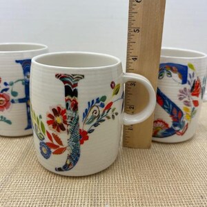 Puede incluir: Tazas de cer&aacute;mica blanca con dise&ntilde;os florales de letras. La letra "J" est&aacute; en el centro, con las letras "H" y "S" visibles en otras tazas. Una regla de madera est&aacute; colocada detr&aacute;s de las tazas.