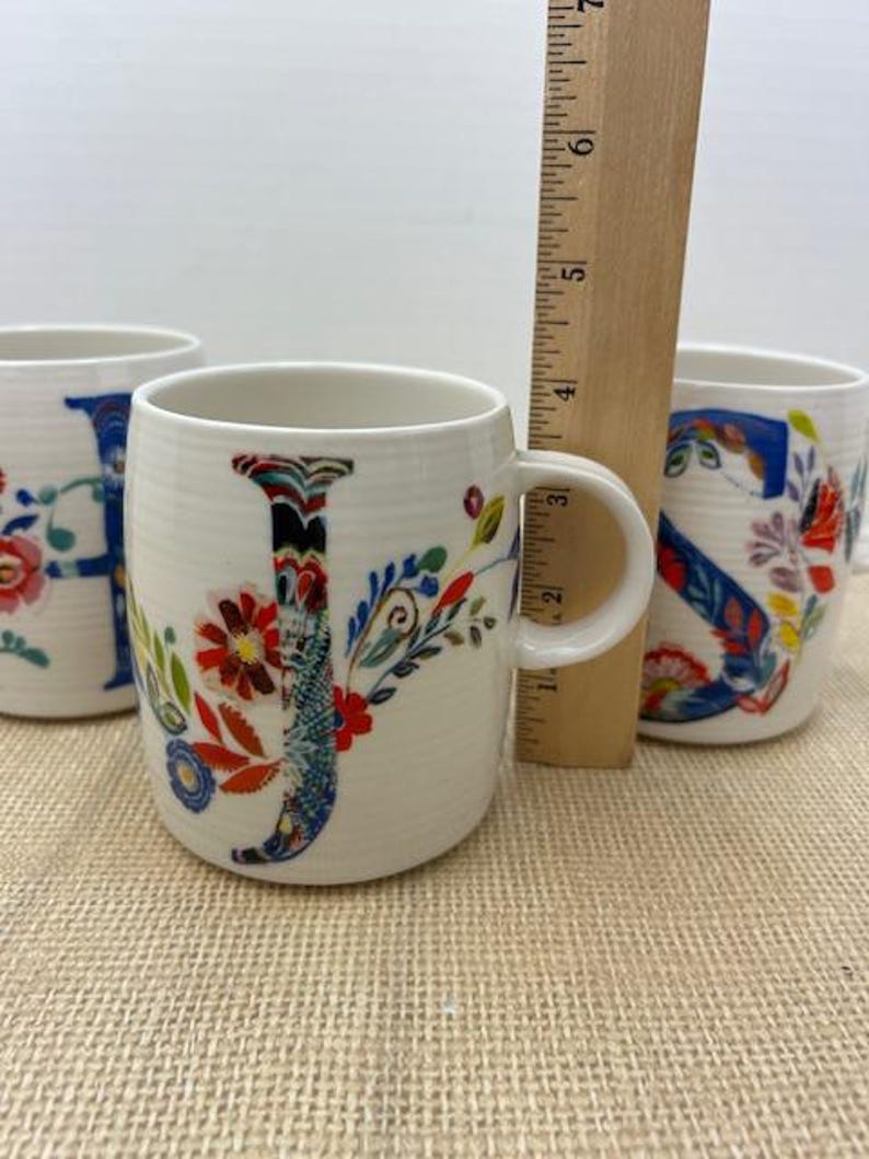 Puede incluir: Tazas de cer&aacute;mica blanca con dise&ntilde;os florales de letras. La letra "J" est&aacute; en el centro, con las letras "H" y "S" visibles en otras tazas. Una regla de madera est&aacute; colocada detr&aacute;s de las tazas.