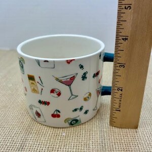 May include: White ceramic mug with a blue handle, decorated with colorful illustrations of cocktails, playing cards, cherries, and billiard balls. A wooden ruler is next to the mug.