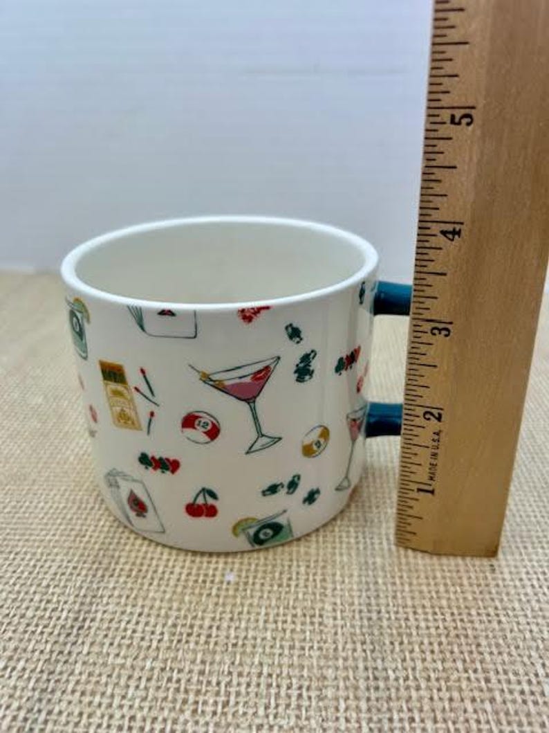 May include: White ceramic mug with a blue handle, decorated with colorful illustrations of cocktails, playing cards, cherries, and billiard balls. A wooden ruler is next to the mug.