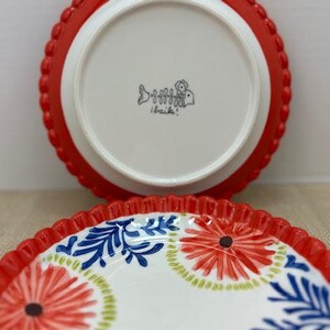 Pu&ograve; includere: Due piatti in ceramica. Uno &egrave; capovolto, mostrando un centro bianco con un bordo smerlato rosso. L'altro piatto mostra una superficie bianca con disegni floreali rossi e motivi a foglie blu. I piatti hanno un aspetto decorativo e artigianale.