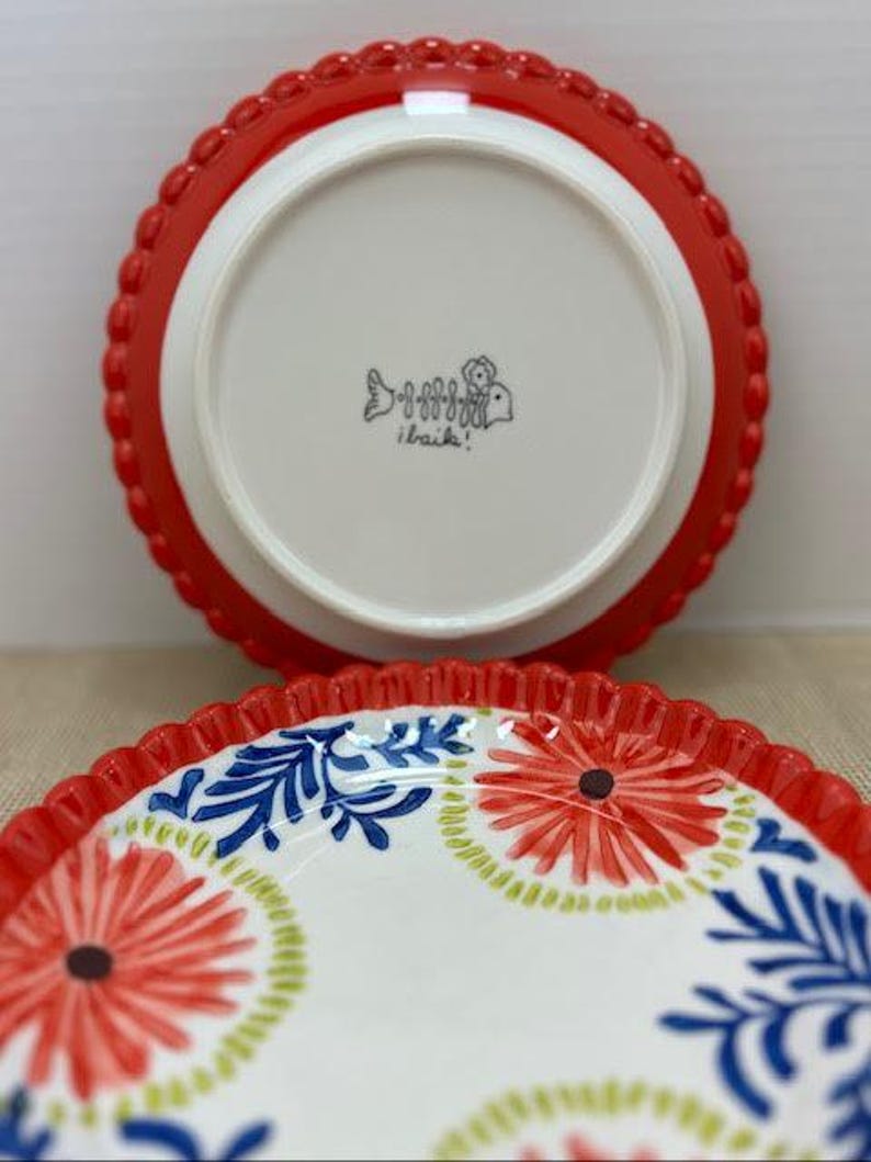 Pu&ograve; includere: Due piatti in ceramica. Uno &egrave; capovolto, mostrando un centro bianco con un bordo smerlato rosso. L'altro piatto mostra una superficie bianca con disegni floreali rossi e motivi a foglie blu. I piatti hanno un aspetto decorativo e artigianale.