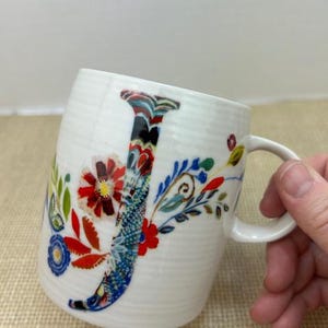 Puede incluir: Taza de cer&aacute;mica blanca con un dise&ntilde;o floral colorido y la letra "J". La taza tiene una forma curva y un asa. El dise&ntilde;o floral incluye detalles en rojo, azul y verde. La taza est&aacute; sujeta por una mano.