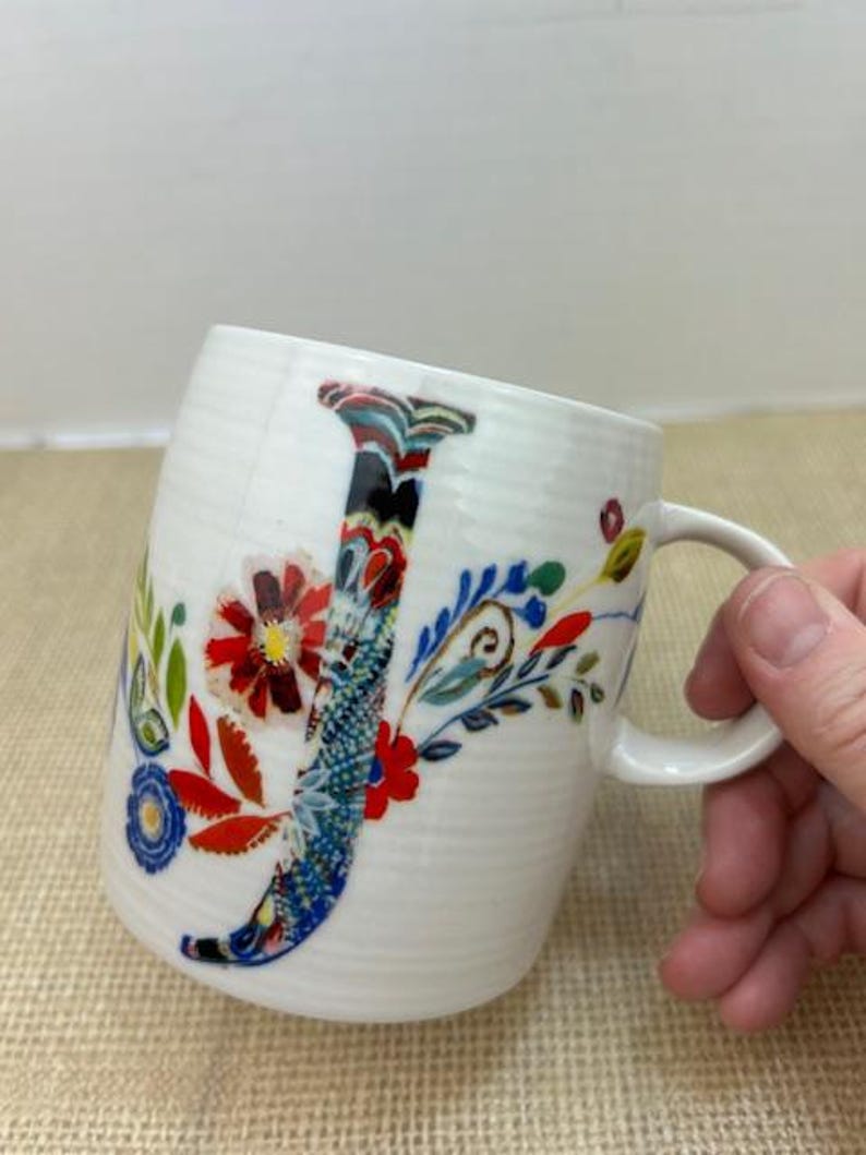 Puede incluir: Taza de cer&aacute;mica blanca con un dise&ntilde;o floral colorido y la letra "J". La taza tiene una forma curva y un asa. El dise&ntilde;o floral incluye detalles en rojo, azul y verde. La taza est&aacute; sujeta por una mano.