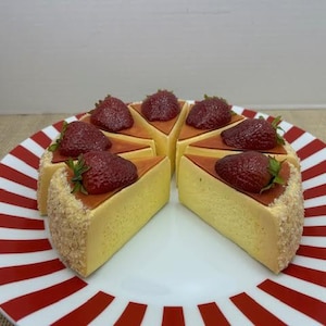 May include: A plate with red and white stripes holds eight slices of cheesecake, each topped with a fresh strawberry. The cheesecake has a golden-yellow filling and a crumbly crust. The strawberries are a deep red.
