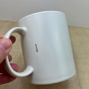 May include: A white ceramic mug with a curved handle. The mug has a cylindrical shape and a smooth surface. The word "&copy;Disney" is printed vertically on the side of the mug. The mug is held by a hand.