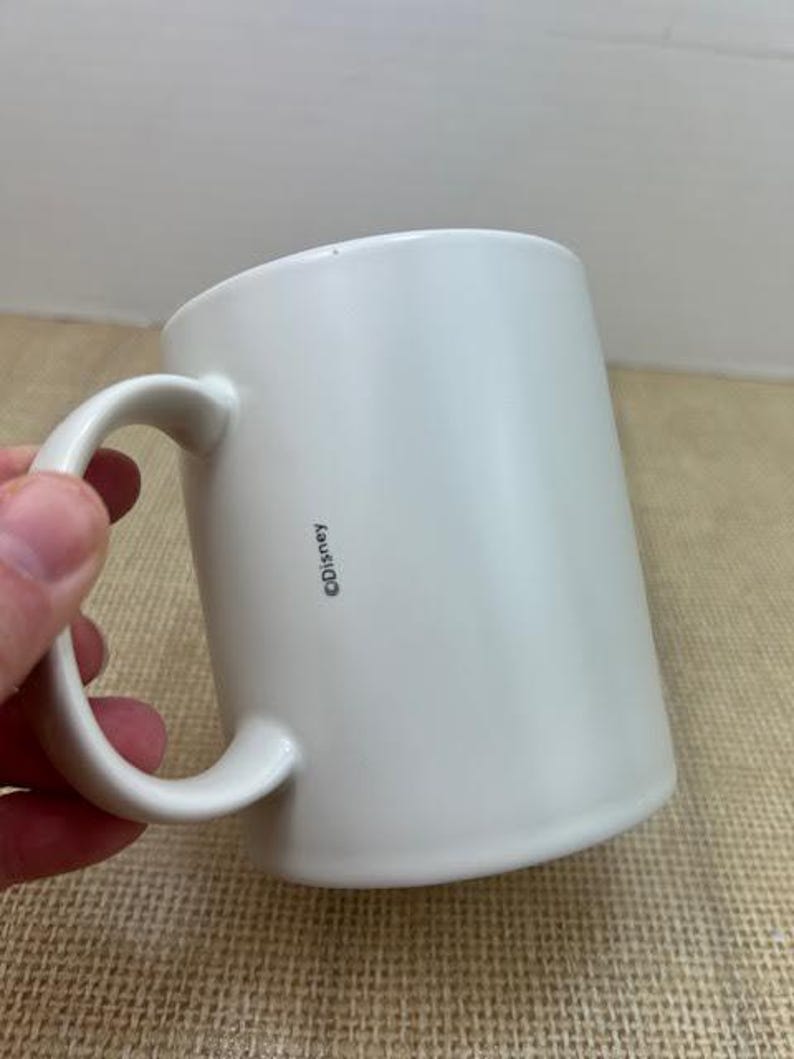 May include: A white ceramic mug with a curved handle. The mug has a cylindrical shape and a smooth surface. The word "&copy;Disney" is printed vertically on the side of the mug. The mug is held by a hand.