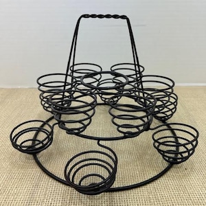 May include: A black metal egg holder with a handle. The holder has six individual compartments for eggs, and a circular base.