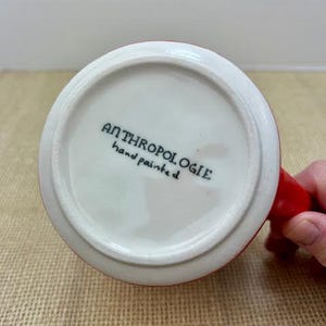 May include: White ceramic mug with a red handle and rim. The bottom of the mug features the text "ANTHROPOLOGIE hand painted" in black lettering. The mug is sitting on a tan surface.