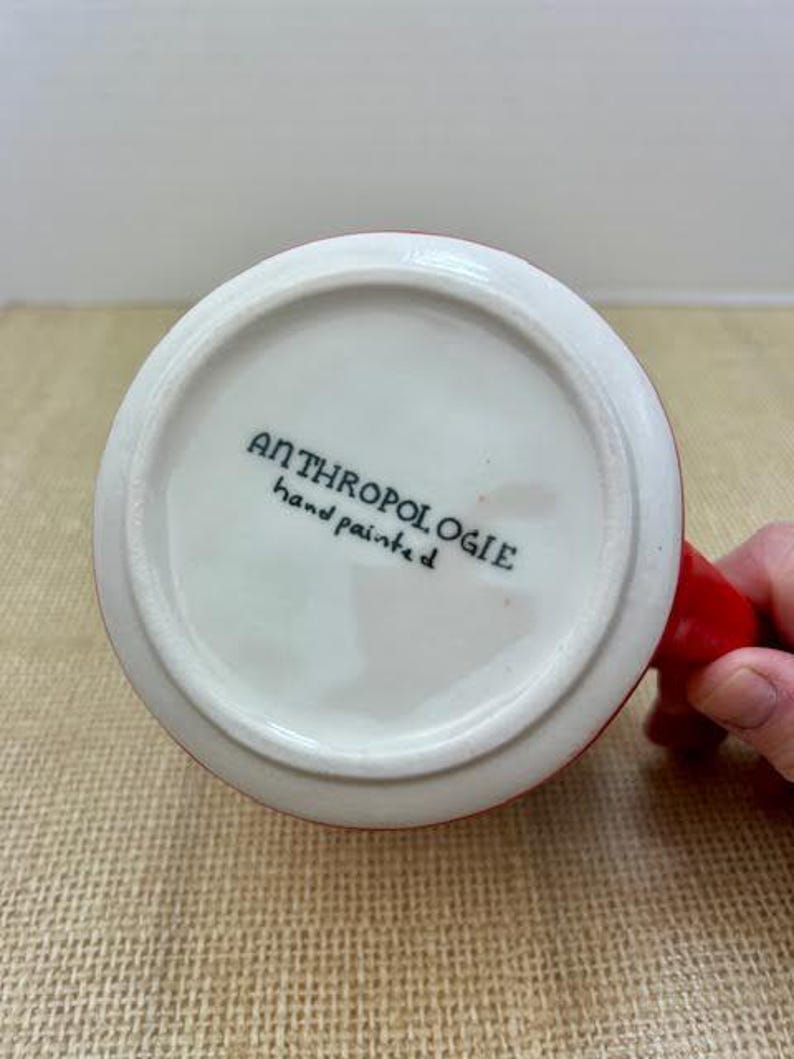 May include: White ceramic mug with a red handle and rim. The bottom of the mug features the text "ANTHROPOLOGIE hand painted" in black lettering. The mug is sitting on a tan surface.