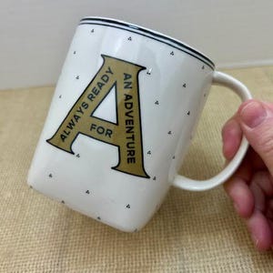 Puede incluir: Taza de cer&aacute;mica blanca con borde negro y peque&ntilde;os lunares negros. La taza presenta una gran letra "A" dorada con las palabras "ALWAYS READY FOR AN ADVENTURE" en texto negro. La taza tiene un asa blanca.