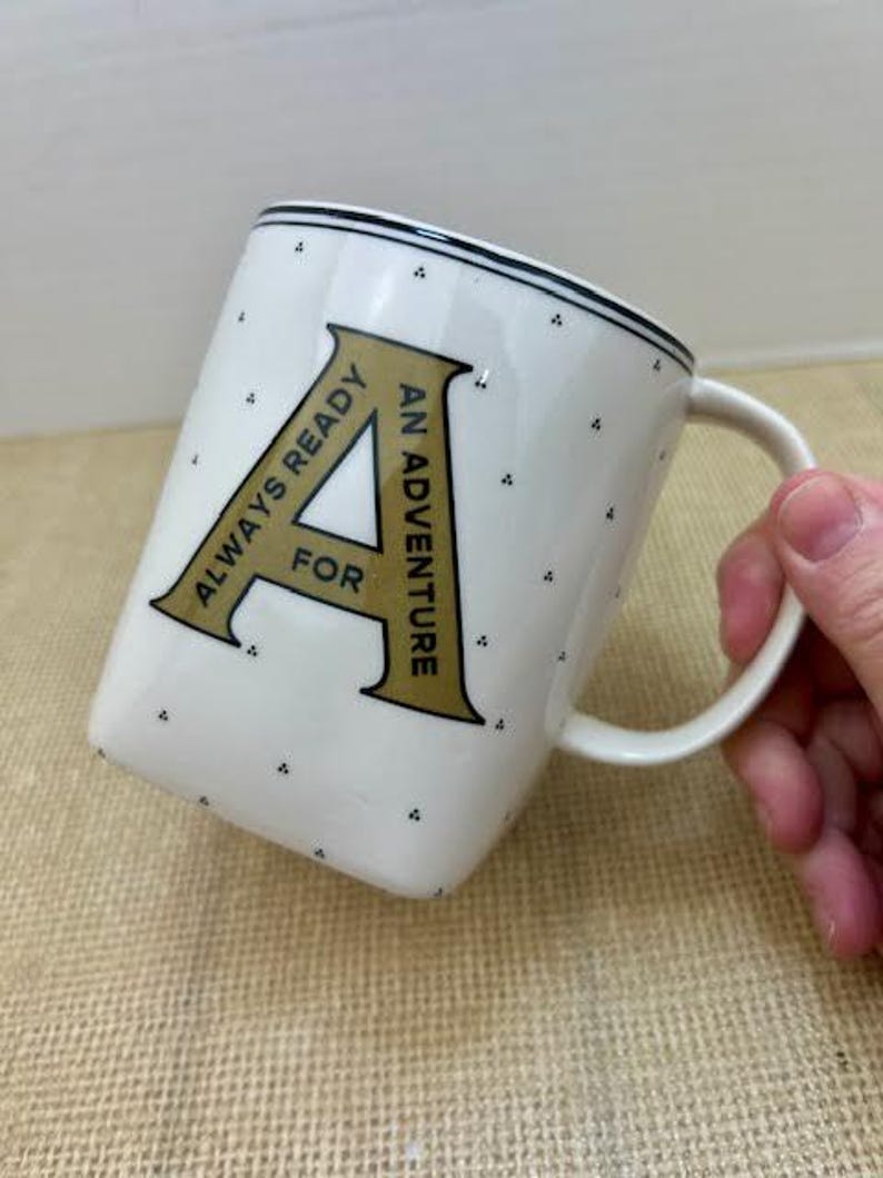 Puede incluir: Taza de cer&aacute;mica blanca con borde negro y peque&ntilde;os lunares negros. La taza presenta una gran letra "A" dorada con las palabras "ALWAYS READY FOR AN ADVENTURE" en texto negro. La taza tiene un asa blanca.
