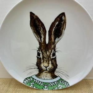 May include: A round white ceramic plate featuring a detailed illustration of a brown hare with long ears. The hare is depicted wearing a green sweater with a white leaf-like pattern around the neckline, centered on the plate surface.