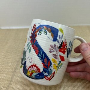 Puede incluir: Taza de cer&aacute;mica blanca con un gran dise&ntilde;o floral de la letra "S". La letra es principalmente azul con detalles florales rojos, amarillos y verdes. La taza tiene una textura acanalada y un asa curva.