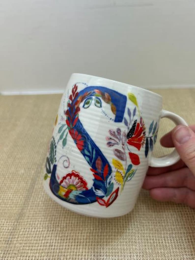 Puede incluir: Taza de cer&aacute;mica blanca con un gran dise&ntilde;o floral de la letra "S". La letra es principalmente azul con detalles florales rojos, amarillos y verdes. La taza tiene una textura acanalada y un asa curva.
