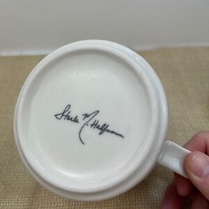 Puede incluir: Taza de cer&aacute;mica blanca con asa, vista desde abajo. La taza presenta una firma en tinta negra. La taza est&aacute; sobre una superficie marr&oacute;n.