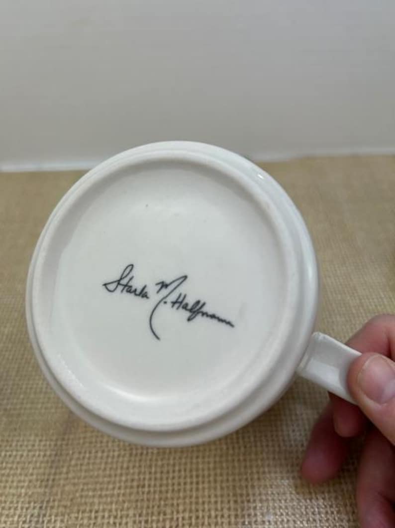 Puede incluir: Taza de cer&aacute;mica blanca con asa, vista desde abajo. La taza presenta una firma en tinta negra. La taza est&aacute; sobre una superficie marr&oacute;n.