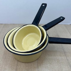 May include: Three nesting enamel saucepans with black rims and handles. The saucepans are yellow and have a vintage look.