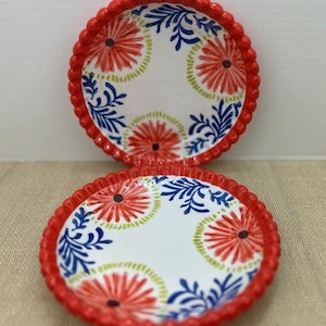 Pu&ograve; includere: Due piatti decorativi in ceramica con bordo smerlato rosso. I piatti hanno uno sfondo bianco con un motivo floreale rosso, blu e verde. I piatti sono rotondi e sembrano piccoli.