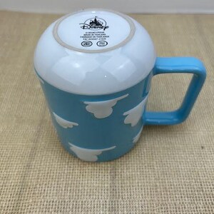 Op de afbeelding: Een lichtblauwe keramische mok met een witte rand en witte wolkontwerpen. De mok heeft het Disney-logo en de tekst "&copy; Disney Pixar Gemaakt in Thailand  Fabriqu&eacute; en Tha&iuml;lande  FAC-403547-27020"