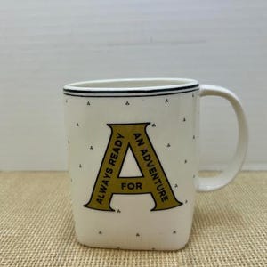 Puede incluir: Taza de cer&aacute;mica color crema con una gran letra "A" dorada y las palabras "ALWAYS READY FOR AN ADVENTURE". La taza tiene un borde punteado negro y un asa blanca. Peque&ntilde;os puntos negros est&aacute;n dispersos por la superficie de la taza.