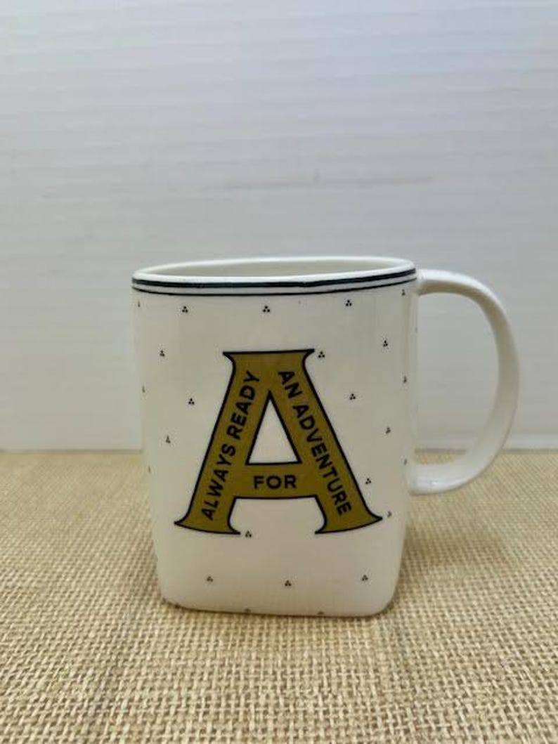 Puede incluir: Taza de cer&aacute;mica color crema con una gran letra "A" dorada y las palabras "ALWAYS READY FOR AN ADVENTURE". La taza tiene un borde punteado negro y un asa blanca. Peque&ntilde;os puntos negros est&aacute;n dispersos por la superficie de la taza.