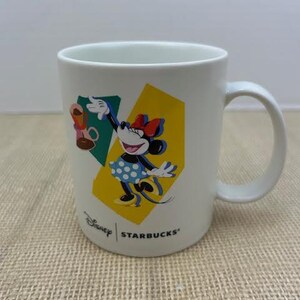 May include: White ceramic mug featuring Minnie Mouse in a polka dot dress, with a red bow, and a yellow and green geometric background. The mug has the Disney and Starbucks logos.