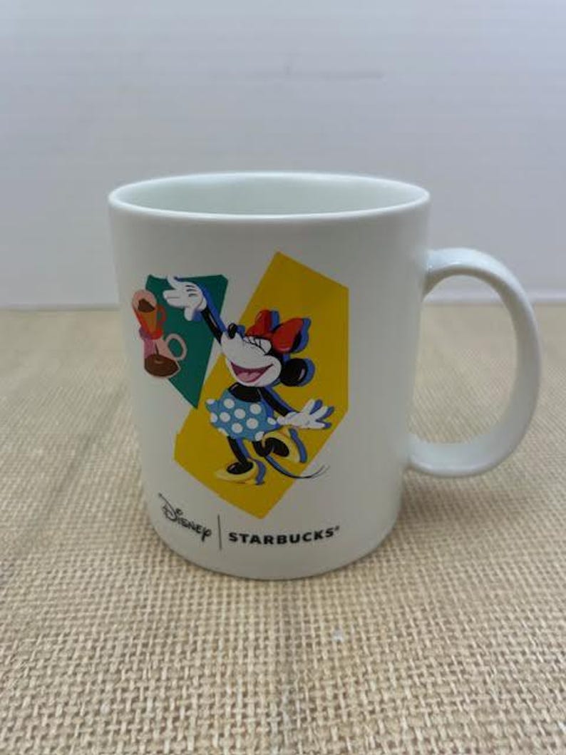 May include: White ceramic mug featuring Minnie Mouse in a polka dot dress, with a red bow, and a yellow and green geometric background. The mug has the Disney and Starbucks logos.