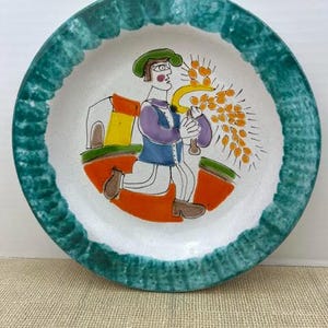 May include: A round ceramic plate with a textured, dark green rim and a white center. The center features a hand-painted illustration of a figure in a blue vest and white pants harvesting orange wheat with a yellow sickle near a small building.