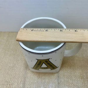 Puede incluir: Taza de cer&aacute;mica blanca con borde y asa negros. La taza presenta una gran letra "A" dorada con el texto "ALWAYS READY AN ADVENTURE". Una regla de madera se coloca en la parte superior de la taza.