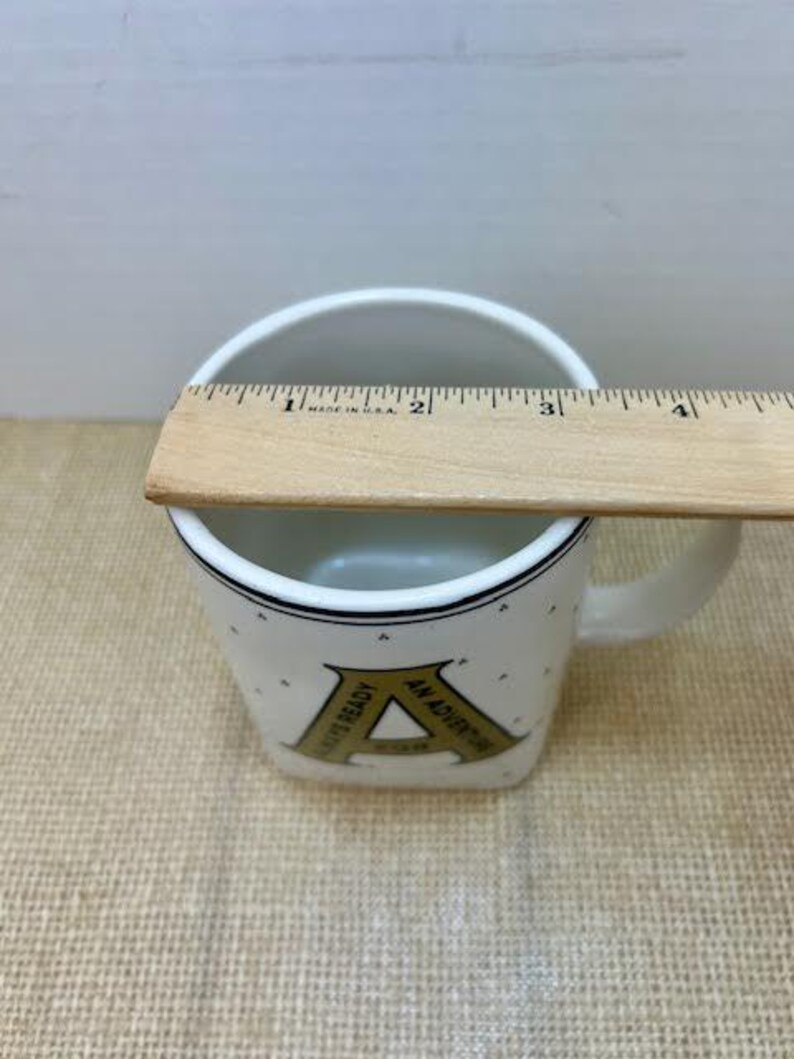 Puede incluir: Taza de cer&aacute;mica blanca con borde y asa negros. La taza presenta una gran letra "A" dorada con el texto "ALWAYS READY AN ADVENTURE". Una regla de madera se coloca en la parte superior de la taza.