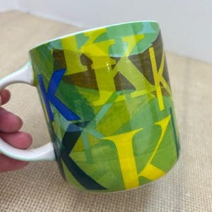 May include: A ceramic coffee mug with a white handle and rim. The mug features a green background with overlapping yellow, blue, and dark green letter "K" designs. The mug is on a tan surface.