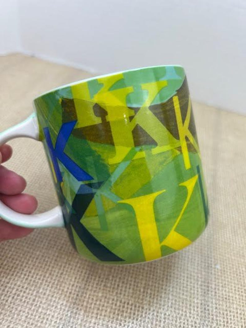 May include: A ceramic coffee mug with a white handle and rim. The mug features a green background with overlapping yellow, blue, and dark green letter "K" designs. The mug is on a tan surface.