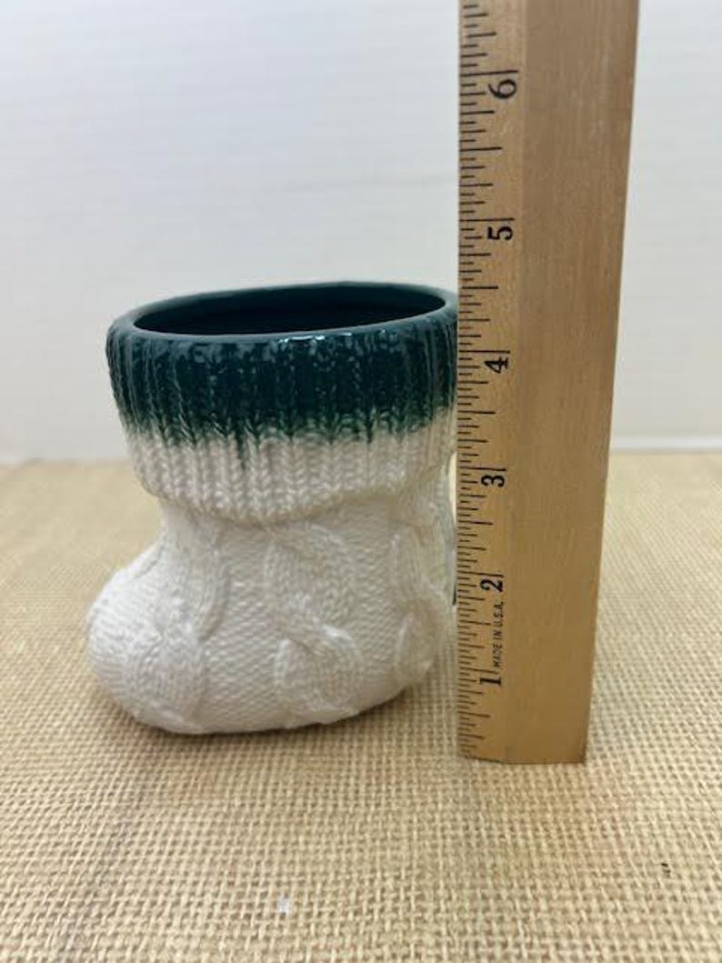 Pu&ograve; includere: Un piccolo vaso in ceramica a forma di calzino. La parte superiore &egrave; verde scuro, che sfuma nel bianco con un motivo a trecce. Un righello di legno misura circa 13 cm.