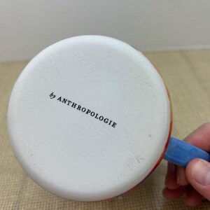 May include: White ceramic mug with a blue handle and colorful accents. The bottom of the mug is visible, with the text "by ANTHROPOLOGIE" printed in black.