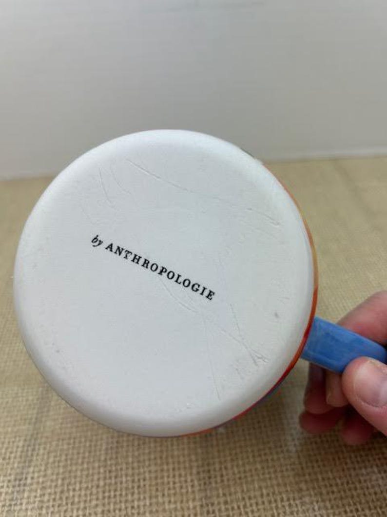 May include: White ceramic mug with a blue handle and colorful accents. The bottom of the mug is visible, with the text "by ANTHROPOLOGIE" printed in black.