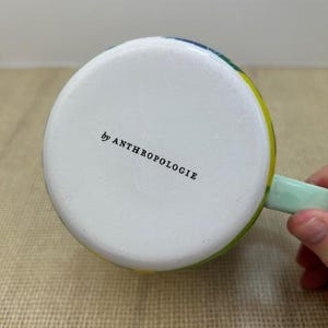 May include: A white ceramic mug with a colorful rim and a light green handle. The bottom of the mug has the text "by ANTHROPOLOGIE" printed in black.