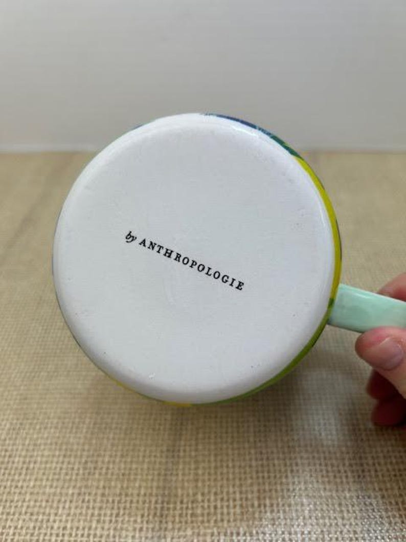 May include: A white ceramic mug with a colorful rim and a light green handle. The bottom of the mug has the text "by ANTHROPOLOGIE" printed in black.