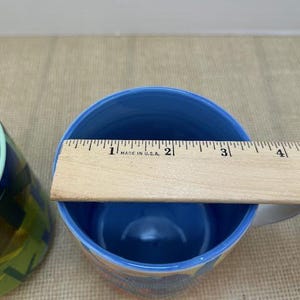 May include: A blue ceramic mug with a light blue interior. A wooden ruler is placed across the top of the mug. The mug has a white handle and a colorful design on the exterior. The ruler measures approximately 3 inches across the top of the mug.