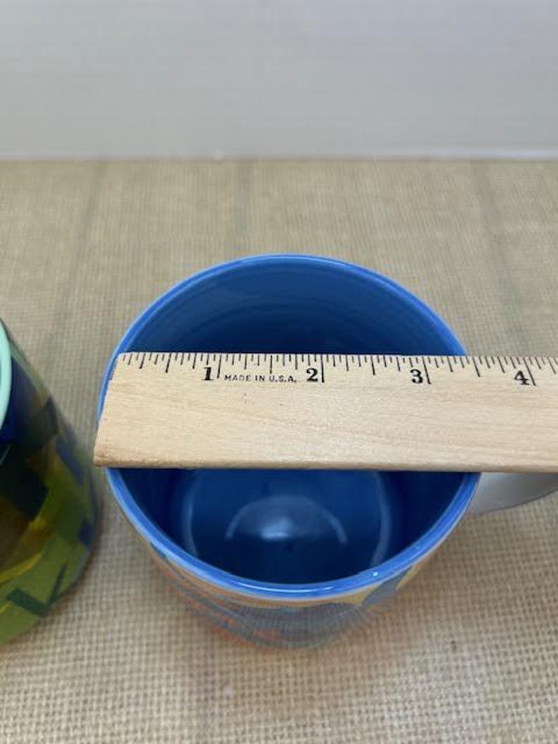 May include: A blue ceramic mug with a light blue interior. A wooden ruler is placed across the top of the mug. The mug has a white handle and a colorful design on the exterior. The ruler measures approximately 3 inches across the top of the mug.