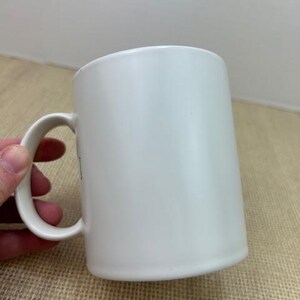 May include: A plain white ceramic mug with a curved handle. The mug has a cylindrical shape and is empty. The mug is held by a hand. The background is a light tan surface.
