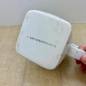 Puede incluir: Taza cuadrada blanca con asa, con peque&ntilde;os puntos negros y el texto "by ANTHROPOLOGIE" en la parte inferior. La taza est&aacute; ligeramente desgastada. El asa es blanca con puntos negros.