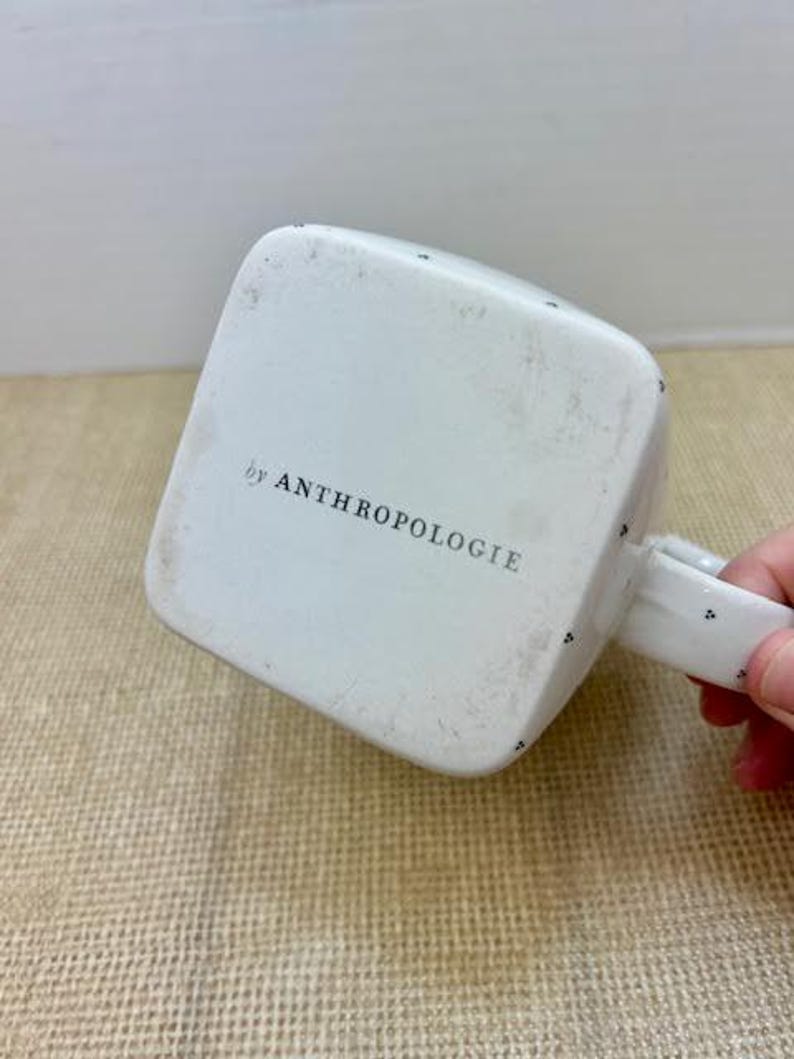 Puede incluir: Taza cuadrada blanca con asa, con peque&ntilde;os puntos negros y el texto "by ANTHROPOLOGIE" en la parte inferior. La taza est&aacute; ligeramente desgastada. El asa es blanca con puntos negros.