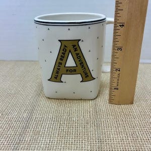 Puede incluir: Taza de cer&aacute;mica blanca con ribete negro y la letra "A" dorada, con las palabras "ALWAYS READY FOR AN ADVENTURE". La taza tiene peque&ntilde;os puntos negros y una regla de madera al lado.